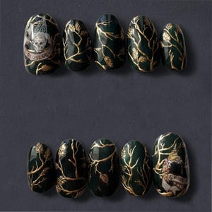 Può includere: Nail art verde scuro con disegni di foglie e rami dorati. Alcune unghie presentano disegni di teschi e banner con i nomi "REGULUS" e "SIRIUS". Le unghie sono di forma ovale e su uno sfondo grigio scuro.