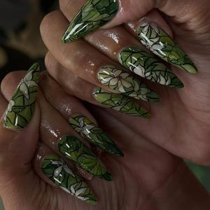 Puede incluir: Uñas largas y puntiagudas con un diseño floral verde y blanco. Las uñas presentan una base verde con patrones de flores y hojas blancas, delineados en negro. El diseño se aplica a cada uña, creando un aspecto cohesivo.