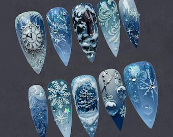 Puede incluir: Un conjunto de diez uñas acrílicas largas y puntiagudas con diseños de invierno. Cada uña presenta un diseño diferente, incluyendo copos de nieve, esferas de reloj y paisajes invernales, en tonos azules, blancos y plateados. Las uñas están decoradas con purpurina y elementos 3D.