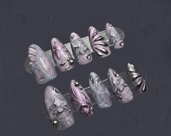 May include: A set of ten artificial nails in various designs. The nails are primarily lavender and silver, with floral and abstract patterns. Some nails feature 3D embellishments, and all have a glossy finish.