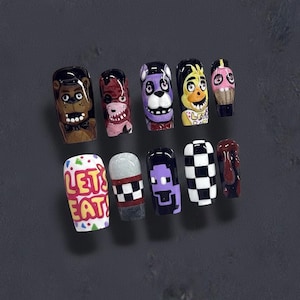 May include: A collection of ten artificial nails, each decorated with a unique design. The designs feature cartoon characters, chequered patterns, and the text "LET'S EAT!" in red and white. The nails are in various colours, including brown, purple, and black.