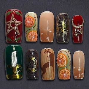 May include: A set of ten artificial nails with various designs. The nails feature a range of colors, including red, green, and nude. Designs include a star, orange slices, a candle, a deer, and gold accents. The nails are arranged on a dark gray surface.