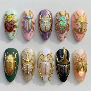 May include: Ten almond-shaped nail art tips featuring three-dimensional gold-toned beetle embellishments. Each tip displays a unique base color, including speckled pastels, glitter, solid green, and black, with beetles featuring opal, floral, and metallic finishes.