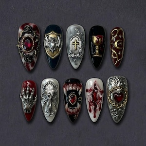 May include: A set of ten long, almond-shaped artificial nails with gothic-inspired designs. Each nail features intricate embellishments, including bats, crosses, chalices, and ornate metalwork with red and black accents. The nails are arranged on a dark gray surface.