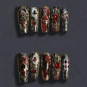 May include: Set of ten coffin-shaped press-on nails. Each nail features a gold metallic design with playing card suit symbols, red glitter, and a large red gem. The nails are arranged on a dark gray surface.