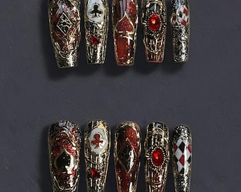 May include: Set of ten coffin-shaped press-on nails. Each nail features a gold metallic design with playing card suit symbols, red glitter, and a large red gem. The nails are arranged on a dark gray surface.