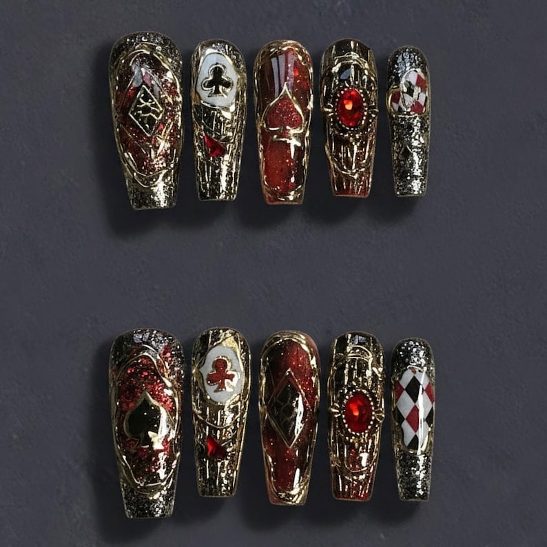 May include: Set of ten coffin-shaped press-on nails. Each nail features a gold metallic design with playing card suit symbols, red glitter, and a large red gem. The nails are arranged on a dark gray surface.