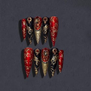Könnte beinhalten: Zehn spitz zulaufende Kunstnägel. Die Nägel sind in Rot, Schwarz und Gold gehalten und weisen geprägte Muster auf. Einige Nägel sind mit Strasssteinen verziert.