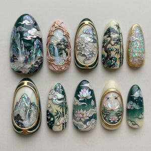 Puede incluir: Un juego de diez uñas postizas en forma de almendra con diseños pintados a mano de montañas, cascadas, flores de loto, bambú y nubes. Las uñas incorporan detalles de concha iridiscente, marcos metálicos dorados y una paleta de colores verde bosque, crema y rosa suave.
