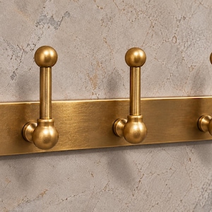 May include: A rectangular brass wall plate featuring three vertical coat hooks with spherical finials. The metal has a brushed finish and is secured to the surface by two visible screws, one on each end of the horizontal mounting bar.