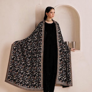 May include: A black velvet long-sleeved tunic and trousers set paired with a large black shawl featuring an intricate, multi-colored floral pattern and a decorative border. The shawl is draped over the shoulders, showcasing the detailed embroidery.