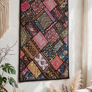 Op de afbeelding: Een rechthoekig wandkleed met een levendige patchwork van geborduurde stoffen vierkanten in tinten rood, roze, geel, blauw en zwart. Het textiel is versierd met kleine reflecterende spiegeltjes, ingewikkeld stikwerk en geometrische patronen.