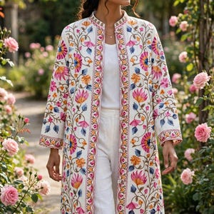 Può includere: Un cappotto color bianco sporco a maniche lunghe con un vivace ricamo floreale multicolore nei toni del rosa, arancione e blu. Il capo presenta un colletto alla coreana, una linea dritta e una chiusura frontale con bottoni su un top bianco.