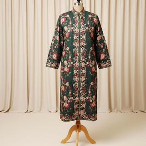 May include: A long, dark green coat with intricate floral embroidery in shades of pink and cream. The coat features a mandarin collar and a button-up front. The sleeves are long, and the coat is displayed on a wooden dress form.