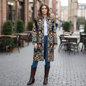 May include: A long, black coat with vibrant floral embroidery, worn by a person. The coat is open, showing a white top and blue jeans. Brown leather boots complete the outfit. The background features a cobbled street and outdoor cafe seating.