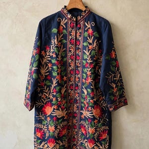 Op de afbeelding: Een marineblauw jasje met lange mouwen, een opstaande kraag en een knoopsluiting aan de voorzijde. Het kledingstuk is versierd met gedetailleerd bloemenborduursel in levendige tinten rood, oranje en groen op de voorzijde en de mouwen.