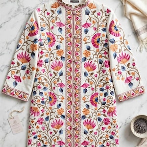 Op de afbeelding: Een witte tuniek met lange mouwen, een opstaande kraag en een centrale sluiting met knopen. Het kledingstuk is versierd met gedetailleerd bloemenborduursel in roze, oranje en blauwe tinten, dat de voorzijde en de mouwen volledig bedekt.