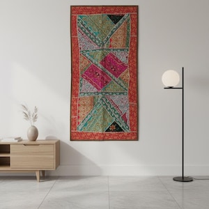 Op de afbeelding: Een rechthoekige wandhanger met een patchwork-ontwerp. Het tapijt heeft een rode rand en een centraal patroon van geometrische vormen in roze, groen en goud. De wandhanger wordt aan een witte muur getoond.