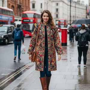 Pode incluir: Uma mulher usando um casaco longo bordado com base preta e padrões florais coloridos. Ela está andando em uma rua molhada em frente a ônibus de dois andares vermelhos e edifícios. Ela está usando botas marrons e uma bolsa marrom.