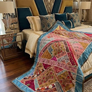 May include: A rectangular patchwork quilt featuring a vibrant array of multi-colored fabric segments in shades of pink, gold, and blue, accented with intricate embroidery, small beads, and a light blue border, draped over a bed with a blue headboard.
