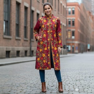 May include: A long, burgundy coat with intricate floral embroidery in yellow and orange. The coat features a mandarin collar and a button-up front. The model is wearing blue jeans and brown boots. The coat is a statement piece.
