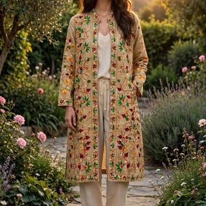 May include: A long, tan, open-front coat featuring intricate, multi-colored floral embroidery in shades of red, green, and yellow. The coat is worn over a simple, light-colored camisole and matching loose-fitting trousers, paired with brown sandals.