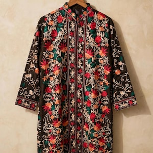 Könnte beinhalten: Eine schwarze Langarmjacke mit einem lebendigen Blumenstickmuster in Rosa-, Orange- und Grüntönen. Die Jacke verfügt über einen Stehkragen, eine mittige Knopfleiste und gerade Ärmel und wird auf einem Holzkleiderbügel präsentiert.