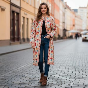 May include: A long, cream-coloured coat with a floral pattern in red, blue, and yellow. The coat is worn over a black top and blue jeans. The person is wearing brown boots and standing on a cobbled street.