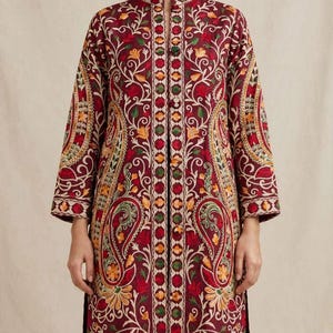 May include: A long-sleeved, knee-length tunic in deep burgundy, featuring intricate, multi-coloured floral and paisley embroidery. The garment has a mandarin collar, a central vertical panel with geometric patterns, and side slits at the hem.
