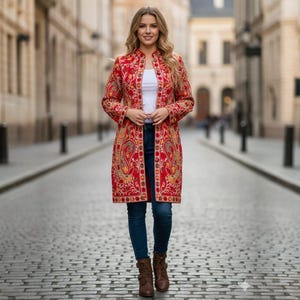 May include: A woman wearing a long, red coat with intricate embroidery. The coat features a mandarin collar and a detailed floral pattern in shades of gold, orange, and green. She is also wearing a white top, blue jeans, and brown boots. The background is a cobblestone street.