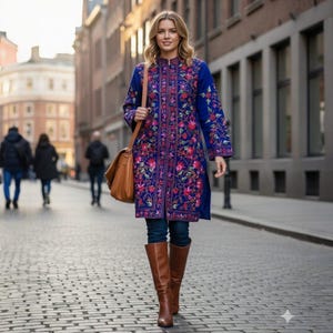 May include: A woman in a long, blue coat with detailed floral embroidery in pink, red, and purple. She carries a brown leather shoulder bag and wears brown boots. The coat is knee-length with long sleeves. The background shows a city street with buildings.