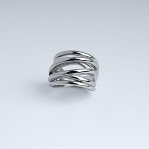 Puede incluir: Un anillo plateado con un diseño tejido. El anillo presenta múltiples bandas de metal que se entrelazan para crear una superficie abierta y texturizada. El anillo está sobre un fondo blanco.
