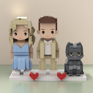 Puede incluir: Un conjunto de figuras estilo Lego que representa a una pareja y un gato. La mujer lleva un vestido azul claro y el hombre un traje beige. Un gato gris está sentado a su lado. Dos corazones rojos están colocados frente a la pareja sobre una base blanca.