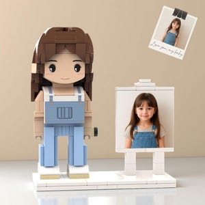 May include: A brick-built figure of a person with brown hair and blue overalls, holding a small object. Beside it, a white easel displays a photo of a young girl. A polaroid photo above reads "Love you, my baby."