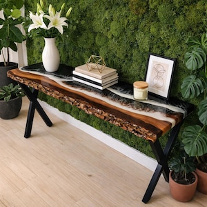 Może przedstawiać: Drewniana ławka z żywą krawędzią i czarnym blatem z żywicy epoksydowej. Ławka ma naturalną krawędź słojów drewna i czarne metalowe nogi. Blat ma czarny, biały i brązowy wzór. Ławka otoczona jest roślinami i zieloną ścianą z mchu.