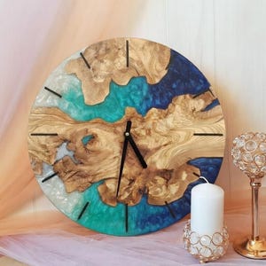 Puede incluir: Un reloj de pared redondo de madera con un diseño único. La esfera del reloj presenta patrones de vetas de madera natural, intercalados con secciones de resina azul, turquesa y blanca. Las manecillas y los marcadores de hora negros completan el diseño. También hay una vela blanca y candelabros decorativos.