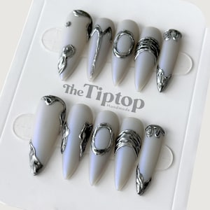 May include: A set of ten long, almond-shaped false nails. Each nail is a pearlescent white with silver metallic accents. Some nails feature a silver swirl design, while others have a silver ring or band. The text "The Tiptop Handmade" is visible.