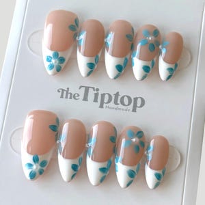 May include: A set of oval false nails. Each nail has a nude base with a white tip, decorated with blue floral patterns. The packaging displays the text "The Tiptop Handmade".
