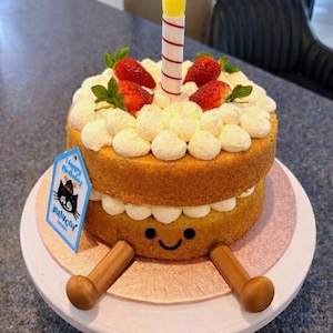 May include: A two-layer birthday cake with white frosting, fresh strawberries, and a lit candle. The cake has a smiling face design and wooden legs. A blue tag with a black cat illustration reads "Happy Birthday".