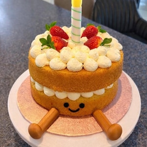May include: A whimsical birthday cake with two layers of yellow sponge cake, decorated with white frosting, fresh strawberries, and a single candle. The bottom layer has a smiling face and wooden legs.