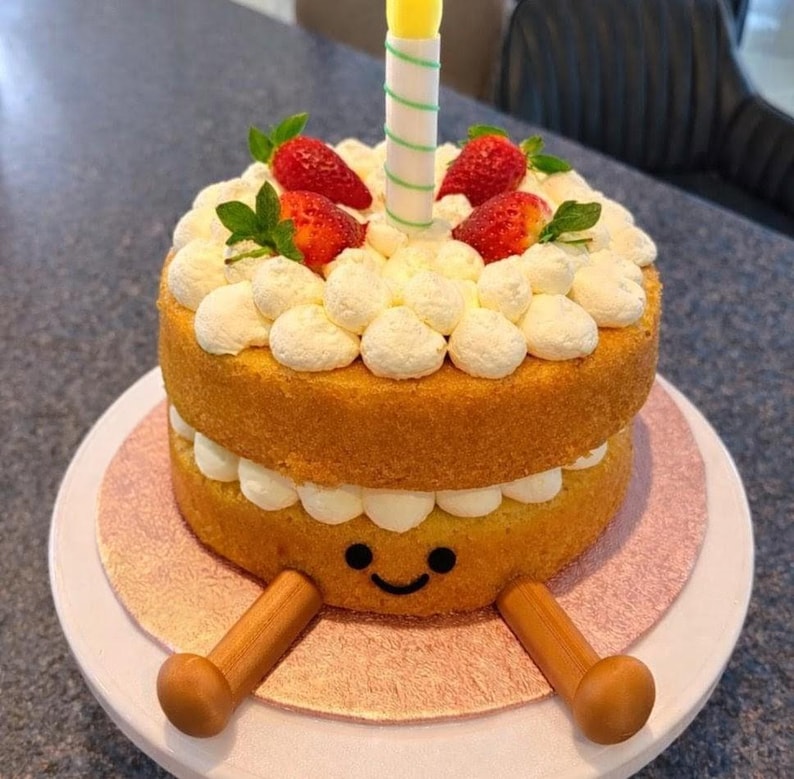 May include: A whimsical birthday cake with two layers of yellow sponge cake, decorated with white frosting, fresh strawberries, and a single candle. The bottom layer has a smiling face and wooden legs.