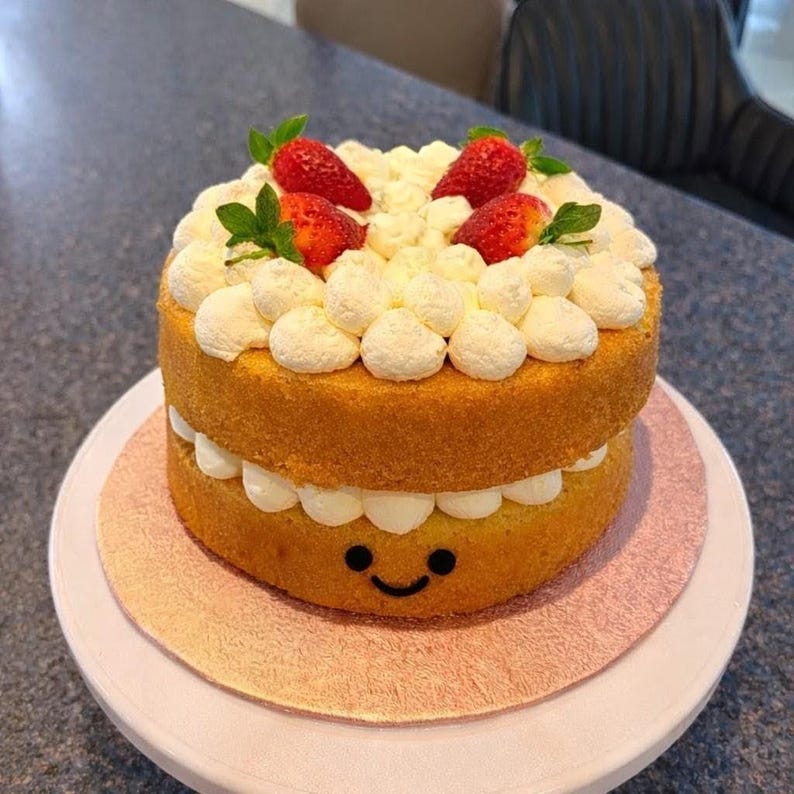 May include: A two-layer cake with white frosting and fresh strawberries. The cake has a smiley face design on the bottom layer. The cake is on a pink cake board and a white cake stand.