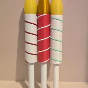 May include: Three decorative candle-shaped objects. Each has a white stem and a cylindrical body wrapped with colored thread. The tops are yellow and gold. The thread colors are red, white, and green.