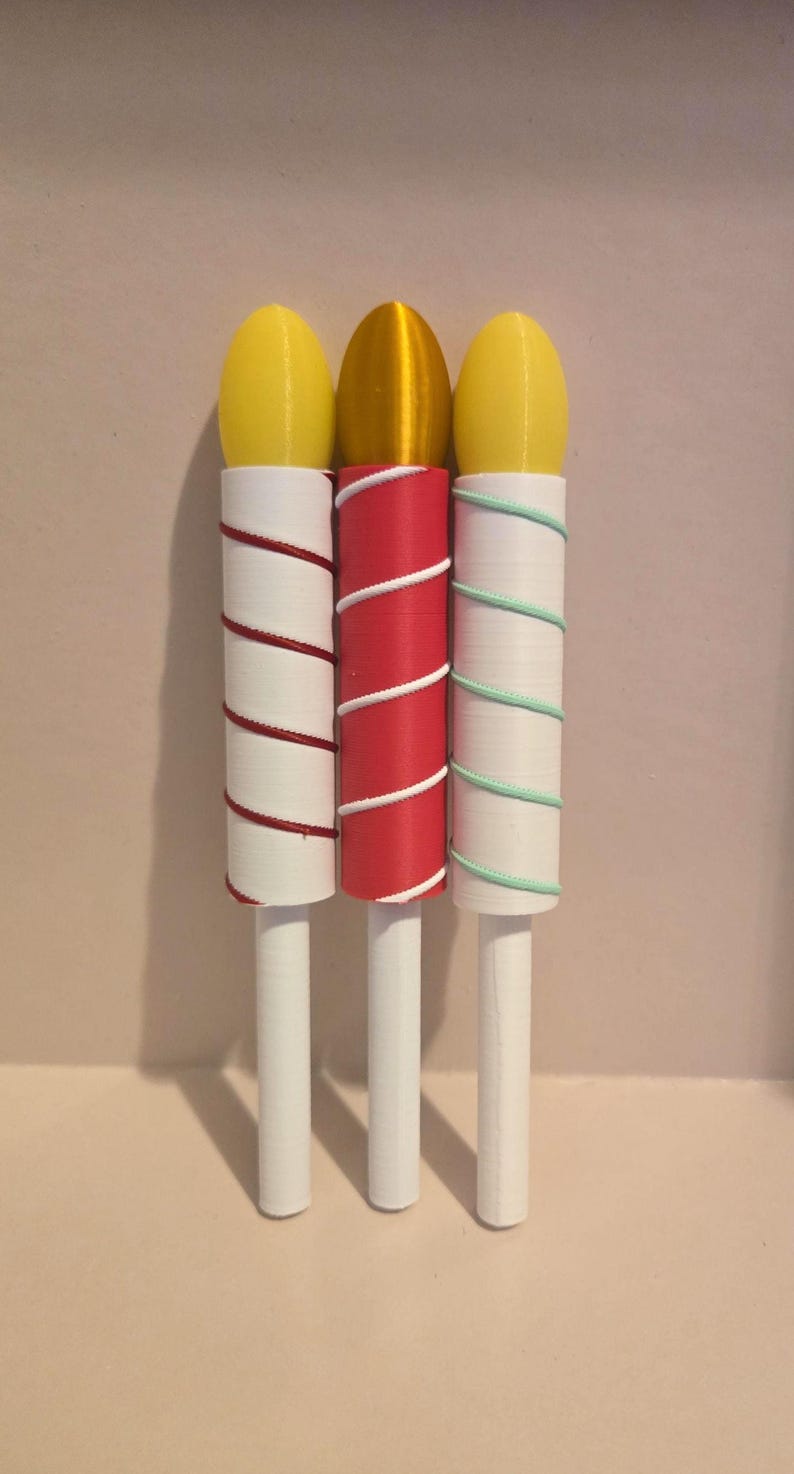 May include: Three decorative candle-shaped objects. Each has a white stem and a cylindrical body wrapped with colored thread. The tops are yellow and gold. The thread colors are red, white, and green.