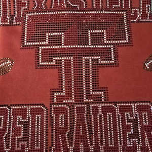 May include: A red fabric garment featuring a large, stylized double T logo and the text RED RAIDERS rendered in sparkling red and white rhinestones. The design is centered on the fabric, with additional rhinestone accents on the sides.