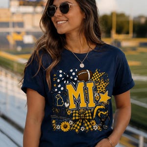 May include: A navy blue short-sleeve t-shirt featuring a graphic print with the letters MI in yellow, a football, a wolverine, sunflowers, and bows in blue and yellow plaid and polka dot patterns, along with a small outline of the state of Michigan.