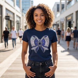 May include: A navy blue short-sleeved t-shirt featuring a large graphic of a blue and white bow with a central star emblem. The bow design has a glittery, textured appearance with a white outline, centered on the chest of the garment.