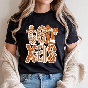 May include: A black short-sleeved t-shirt featuring a large graphic on the front that spells out the word TEXAS in stylised letters. The letters are decorated with orange and white patterns including leopard print, checkers, stripes, stars, and bows.