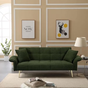 May include: A dark green sofa with two matching cushions. The sofa has gold-coloured legs and is positioned against a beige wall with framed artwork. A lamp with a white shade is on the right.