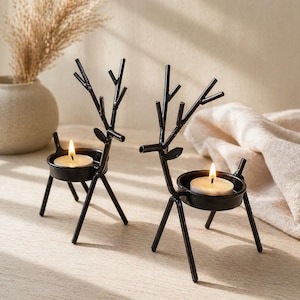 Puede incluir: Dos candelabros de reno de metal negro, cada uno con una vela encendida en un soporte redondo. Los renos tienen cornamentas y se apoyan sobre tres patas. Un jarrón con plantas secas está en el fondo.